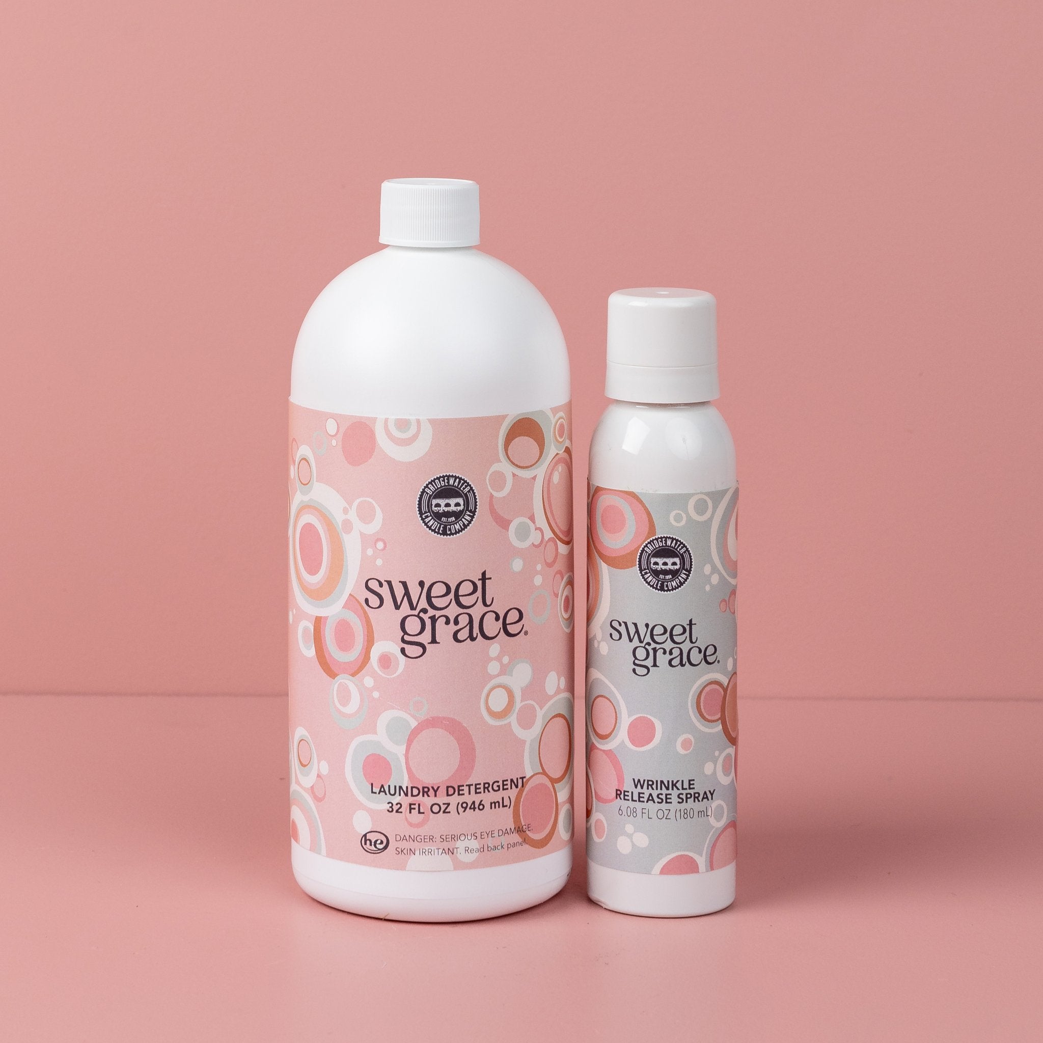 Two pink bottles from the Sweet Grace Laundry Bundle stand on a pink surface, with Sweet Grace Laundry Detergent on the left and Wrinkle Release Spray on the right, both featuring circular patterned labels.