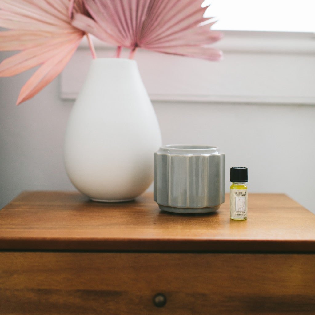 A white vase with pink dried palm leaves, a gray ceramic container, and a Fast Bundle Sweet Grace Home Fragrance Oil 3 Pack Bundle sit on a wooden table against a white wall.