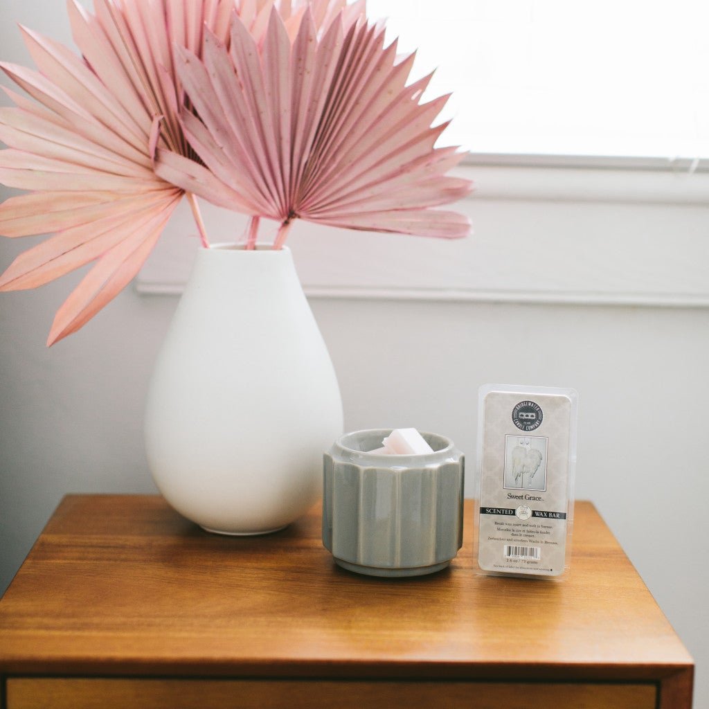A white vase with pink dried palm leaves, a gray wax warmer with soy-blend melts, and a Fast Bundle Sweet Grace Wax Bar 3 Pack Bundle sit on a wooden table by a window, offering flameless fragrance.