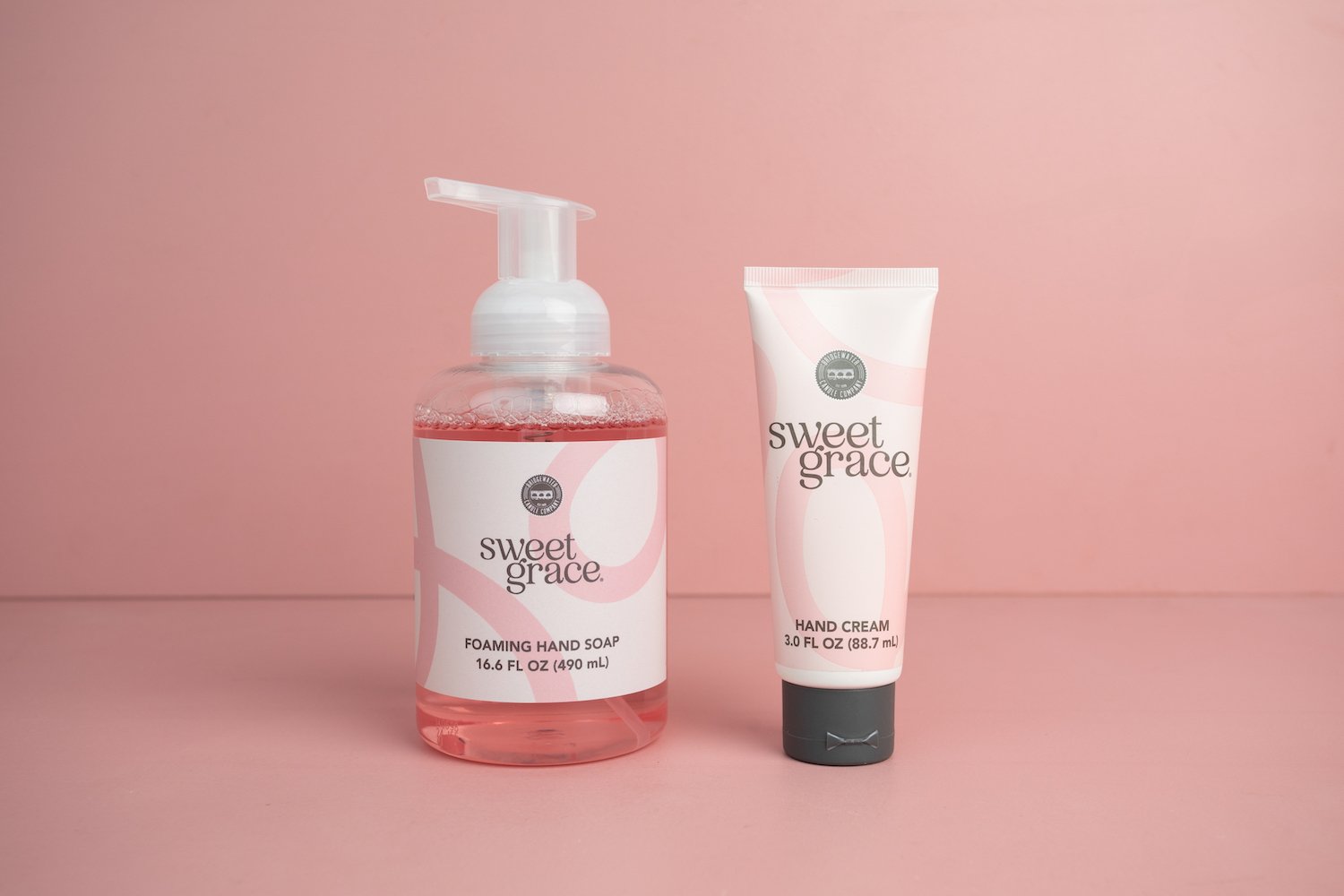 The Sweet Grace Foam & Pamper Bundle, featuring foaming hand soap and hand cream, is displayed side by side on a pink surface with a matching pink background.