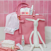 A pink diaper bag sits before a tiled wall, paired with baby essentials and Bridgewater Candle's Sweet Grace Stain Remover (Travel Size) for quick clean-ups on the go.