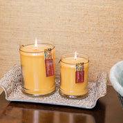 Two lit Bridgewater Candle Autumn Gold Large Jar Candles with brown labels sit on a tray atop a dark wood surface, set against a textured beige background with part of a ceramic bowl visible on the right.