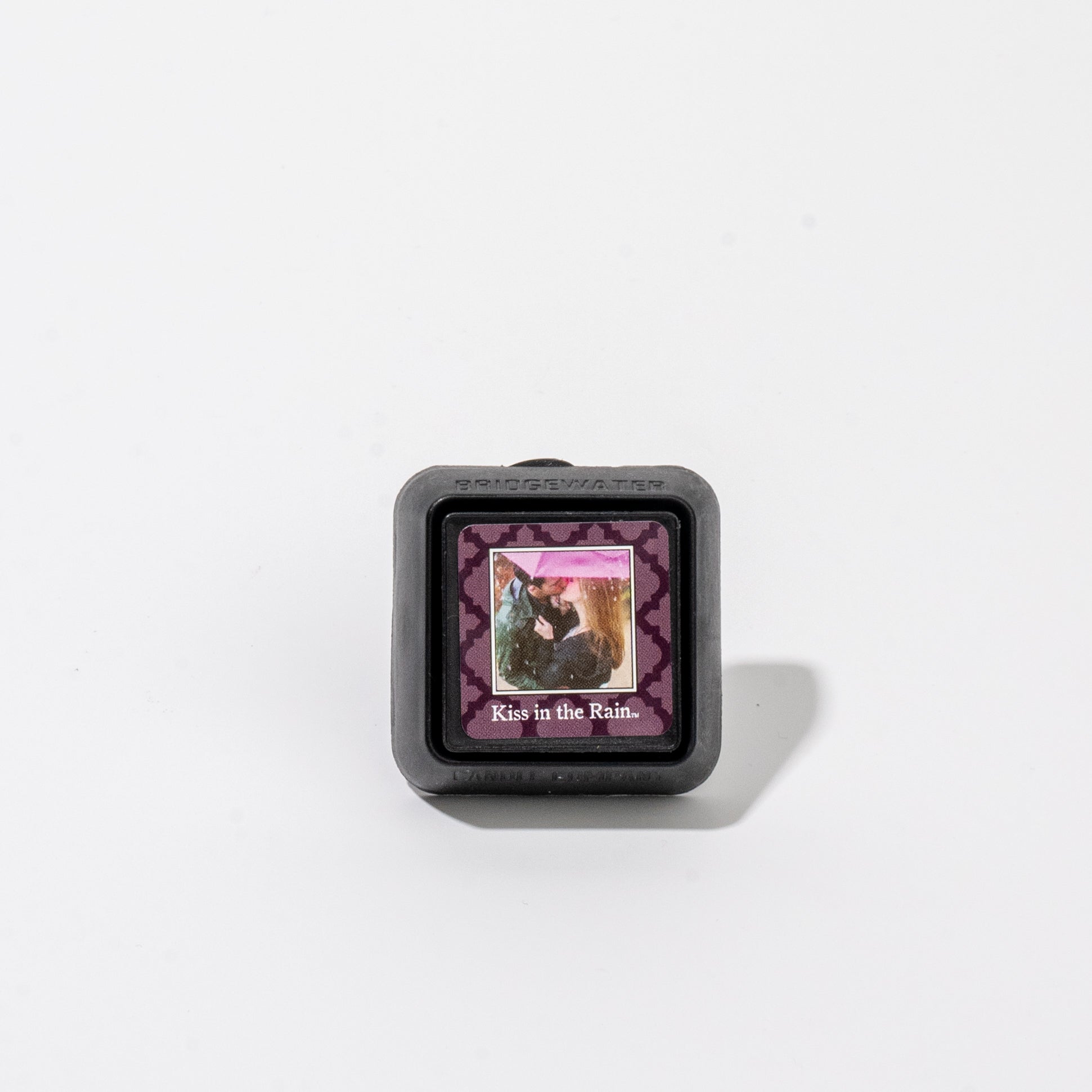 A square black Auto Vent Clip-Kiss in the Rain with a label featuring an illustration of a couple kissing under a pink umbrella and the text Kiss in the Rain.