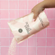 A hand with pink-painted nails pours Bridgewater Candle’s Sweet Grace Soaking Salts from a pastel pink and white bag in front of a pink tiled background.