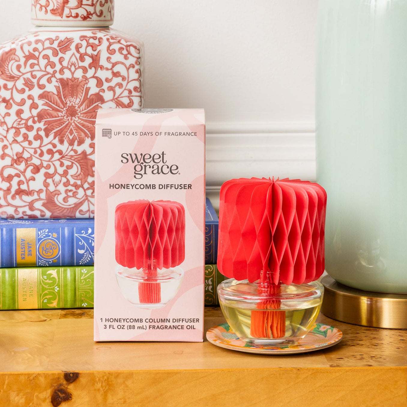 A decorative table with colorful books, a patterned pink jar, the Bridgewater Candle Sweet Grace Honeycomb Diffuser for ongoing fragrance, and a mint green lamp with a multicolored shade set against a crisp white wall.