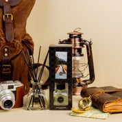 A Bridgewater Candle Solitude Reed Diffuser set with fiber reeds and a scenic box rests on a beige surface beside a vintage camera, brown leather backpack, lantern, compass, and leather-bound notebook for premium home fragrance.