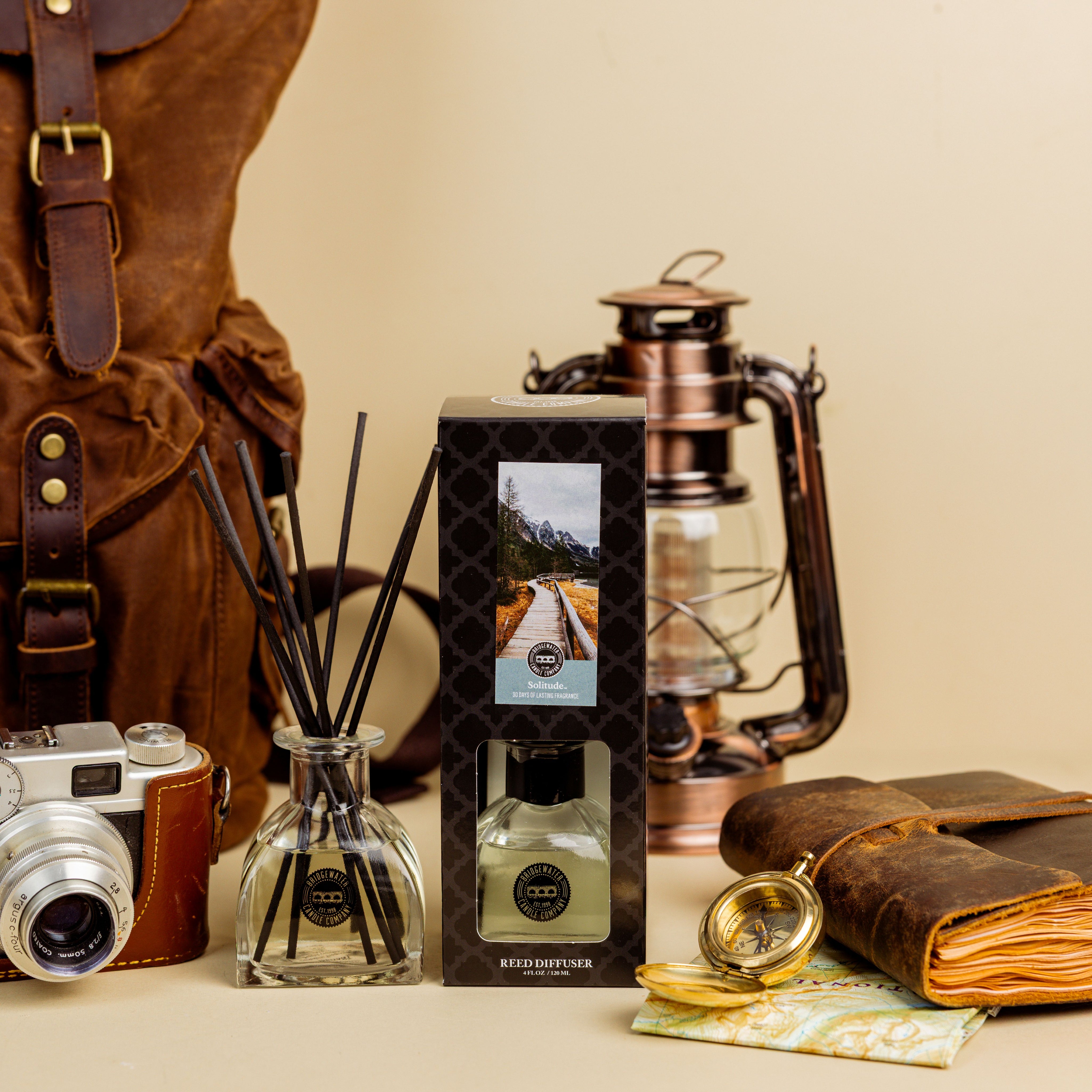 A Bridgewater Candle Solitude Reed Diffuser set with fiber reeds and a scenic box rests on a beige surface beside a vintage camera, brown leather backpack, lantern, compass, and leather-bound notebook for premium home fragrance.