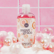 A bottle of Bridgewater Candle Sweet Grace Bubble Bath surrounded by pink and white soap bubbles against a tiled wall.