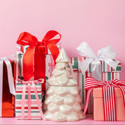 Several colorful gift boxes with red, white, and pink ribbons surround a Bridgewater Candle Sweet Grace Collection Candle #70, crafted from soy-blend wax in a white ceramic Christmas tree, all set against a soft pink background.