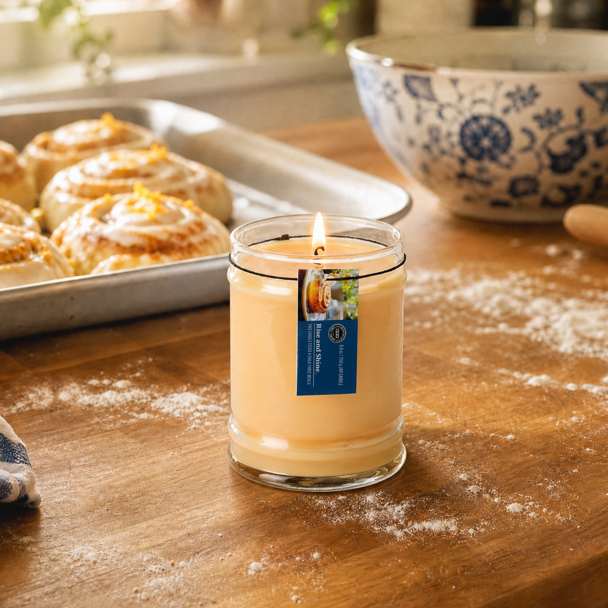 A lit Rise & Shine 8oz Jar Candle by Bridgewater Candle sits on a wooden kitchen counter beside frosted cinnamon rolls and a patterned bowl. Scattered flour adds to the cozy, soy-blend wax aroma.