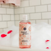 A clear bottle of Bridgewater Candle Sweet Grace Bubble Bath rests in a bubble-filled tub with scattered rose petals, ideal for a moisturizing self-care routine.