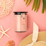The Bridgewater Candle Sunkissed 18oz Large Jar Candle, crafted from soy-blend wax, sits in a glass jar with a black lid on a pink background, surrounded by a starfish, seashell, netting, palm leaves, and a straw hat with a bow.
