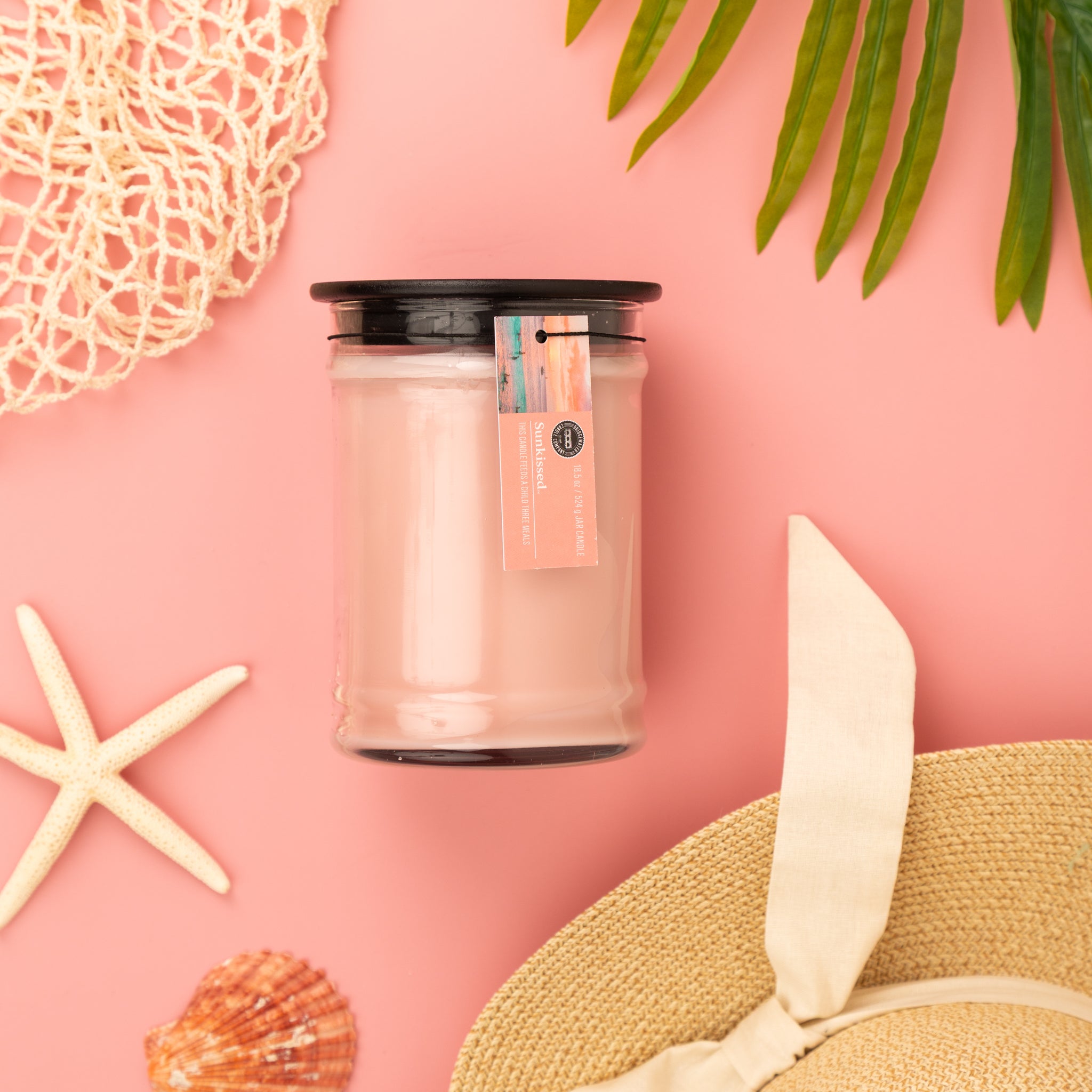 The Bridgewater Candle Sunkissed 18oz Large Jar Candle, crafted from soy-blend wax, sits in a glass jar with a black lid on a pink background, surrounded by a starfish, seashell, netting, palm leaves, and a straw hat with a bow.