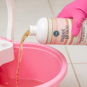 A person wearing a pink glove pours Bridgewater Candle Sweet Grace Cleaning Concentrate | Mop Soap, a fragrant cleaner, into a pink bucket on a tiled floor.