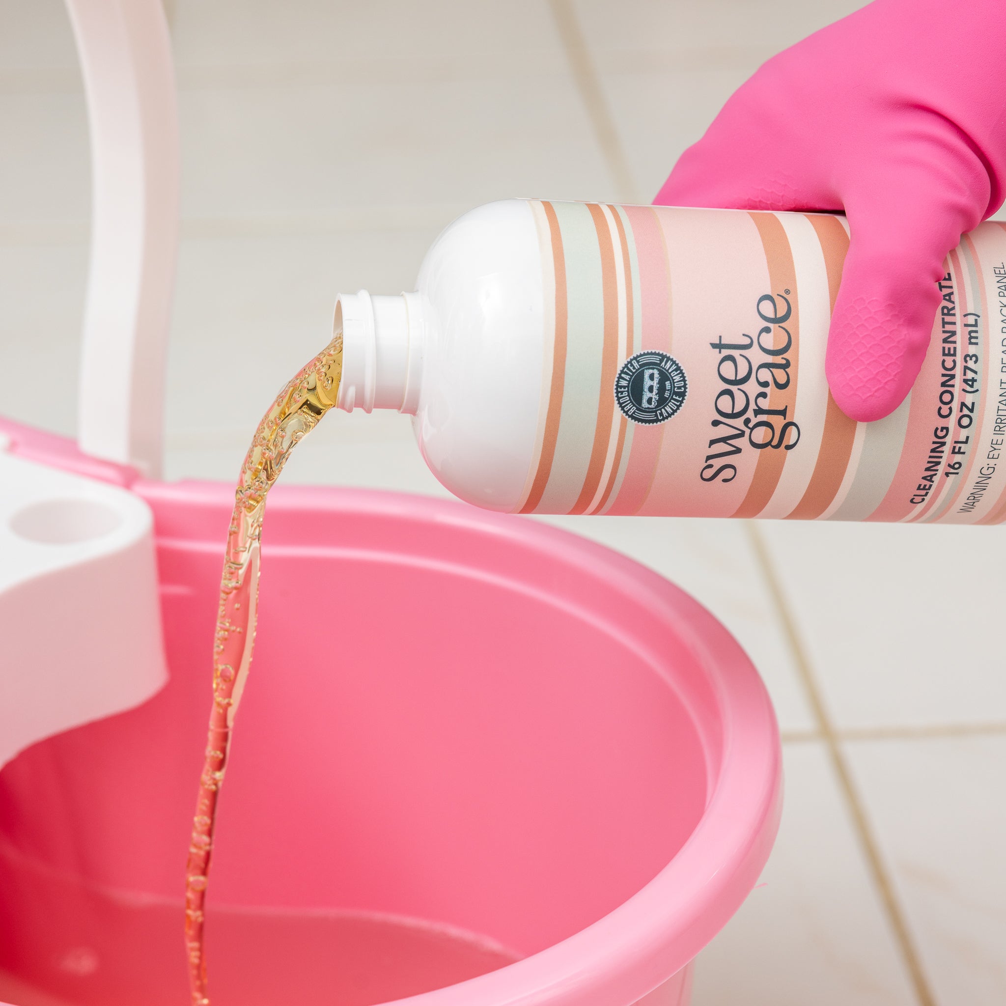 A person wearing a pink glove pours Bridgewater Candle Sweet Grace Cleaning Concentrate | Mop Soap, a fragrant cleaner, into a pink bucket on a tiled floor.
