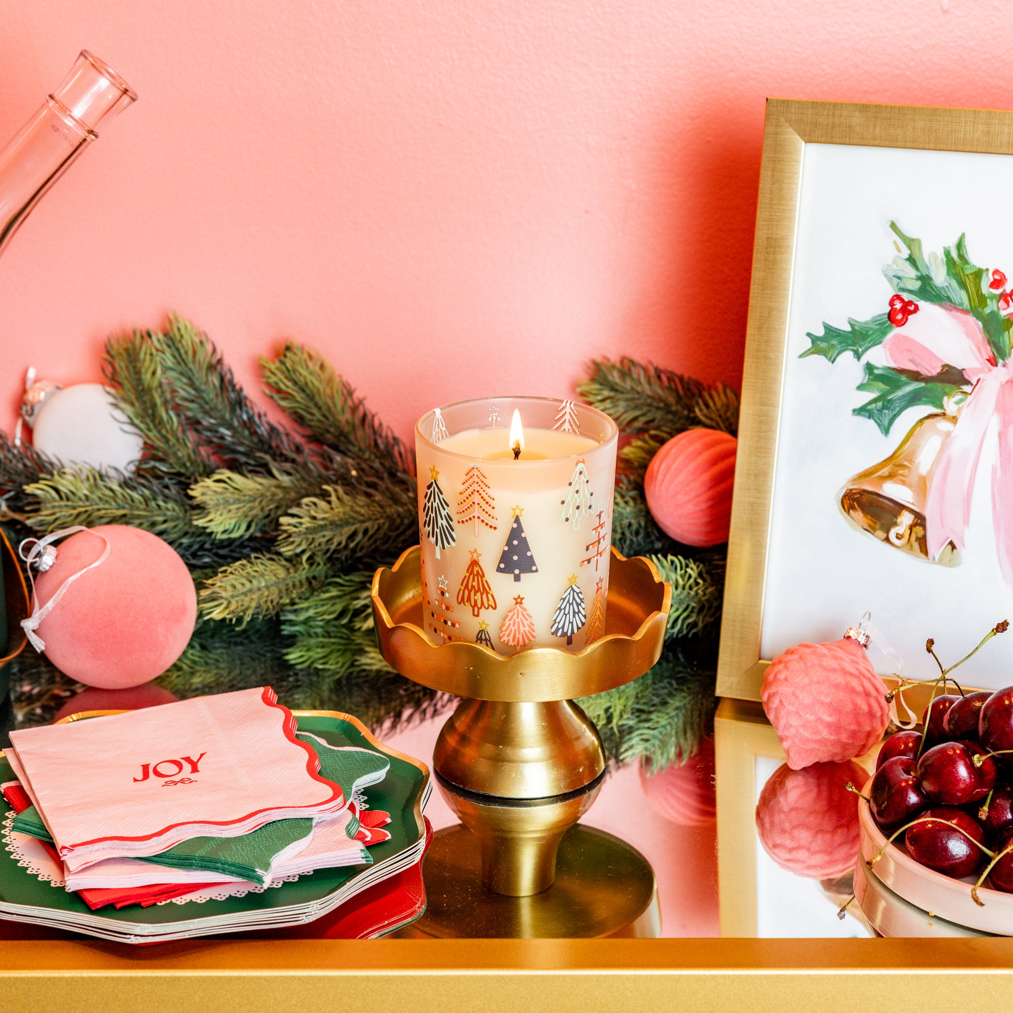 A festive holiday scene highlights the Bridgewater Candle Sweet Grace Holiday Tree Candle as the centerpiece, surrounded by Christmas napkins, ornaments, evergreen branches, a framed bells painting with holly, and cherries on a pink and gold tabletop.