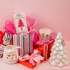 A festive holiday setup with striped and red gift bags, a Santa mug, Bridgewater Candle’s Sweet Grace Holiday Honeycomb Diffuser for lasting fragrance, a nutcracker candle, pearl necklace, white ceramic Christmas tree, and gifts on a pink background.
