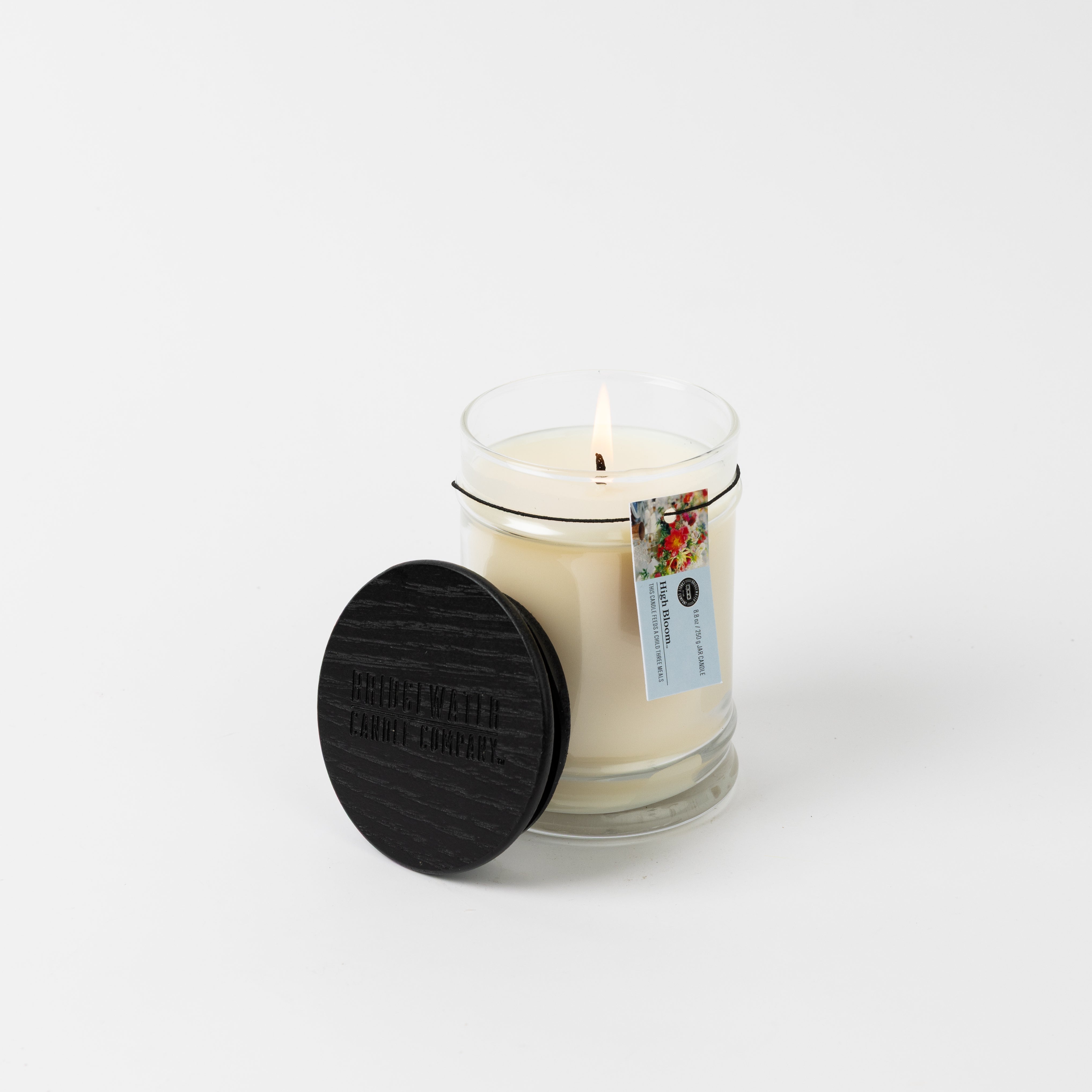 The High Bloom 8oz Small Jar Candle by Bridgewater Candle glows in a clear glass jar with a black wooden lid and floral tag, filled with soy-blend wax. The fragrant candle stands out against a plain white background.