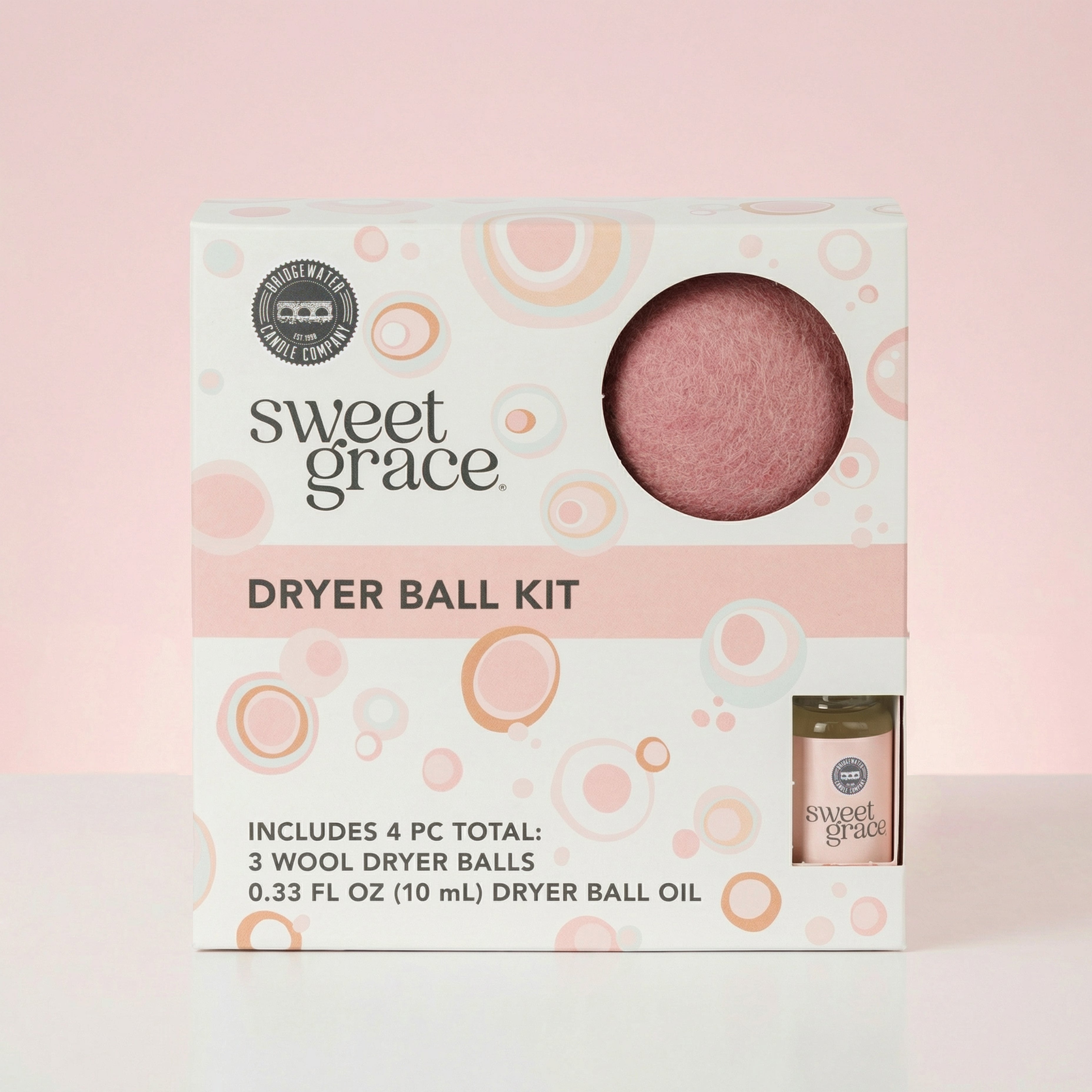A Bridgewater Candle Sweet Grace Dryer Ball Kit box sits against a pink background, displaying 3 wool dryer balls (one visible through a window) and a small bottle of fragranced oil. The white packaging features pink and orange circle designs.