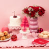 A festive pink table features a coconut cake with cherries, red roses with baby's breath, Christmas ornaments, pastries, red “PEACE” napkins, and a Bridgewater Candle Sweet Grace Holiday Honeycomb Diffuser for lasting fragrance.