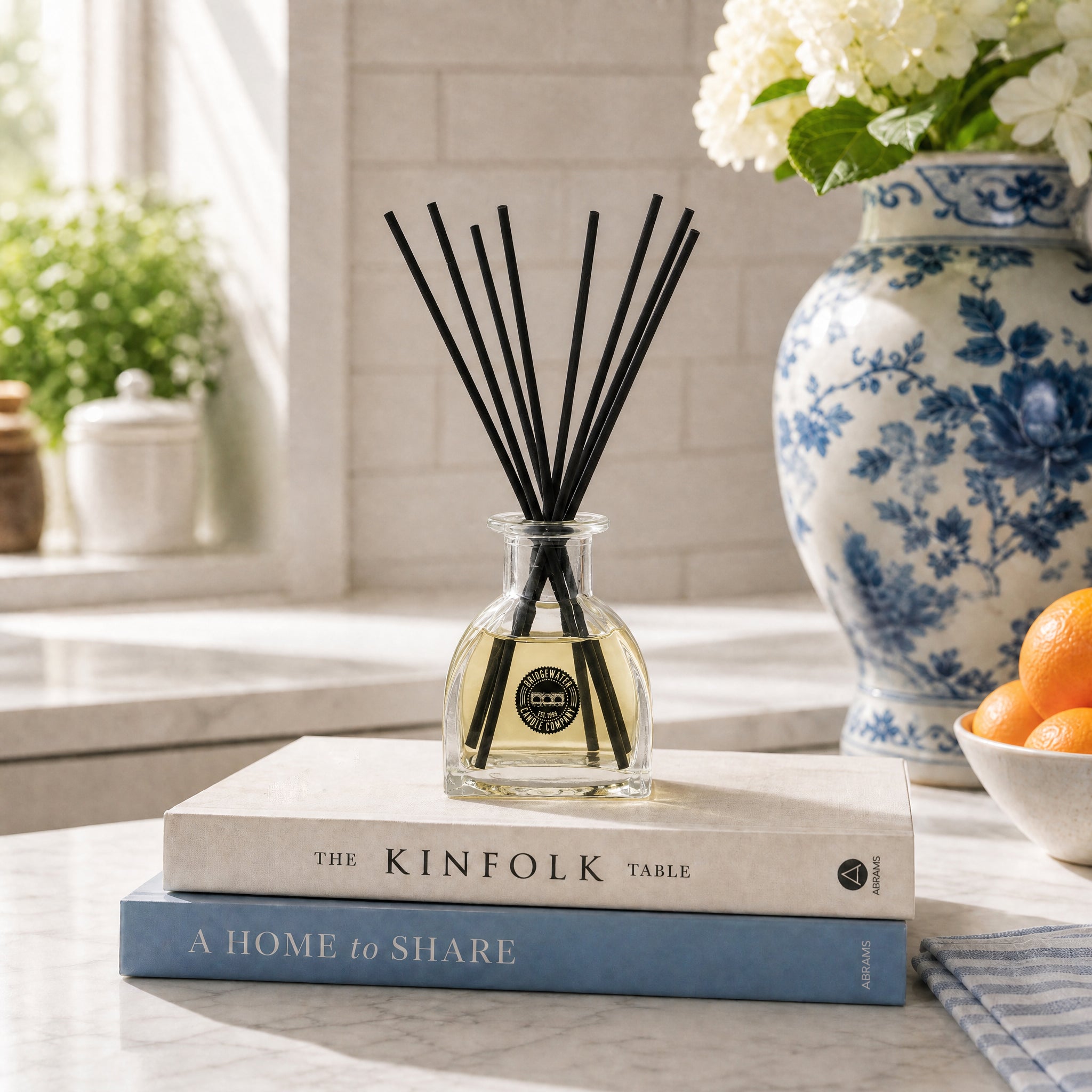 A Bridgewater Candle Rise & Shine Reed Diffuser sits on two stacked books on a sunny kitchen counter. Behind it, a blue and white floral vase with white flowers and a bowl of oranges create an inviting home fragrance scene.