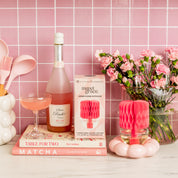 A pink-themed kitchen scene with pink utensils, a bottle and glass of pink Bellini, pink flowers, a Bridgewater Candle Sweet Grace Honeycomb Diffuser for lasting fragrance, and two pink books on a white counter against a pink tiled wall.