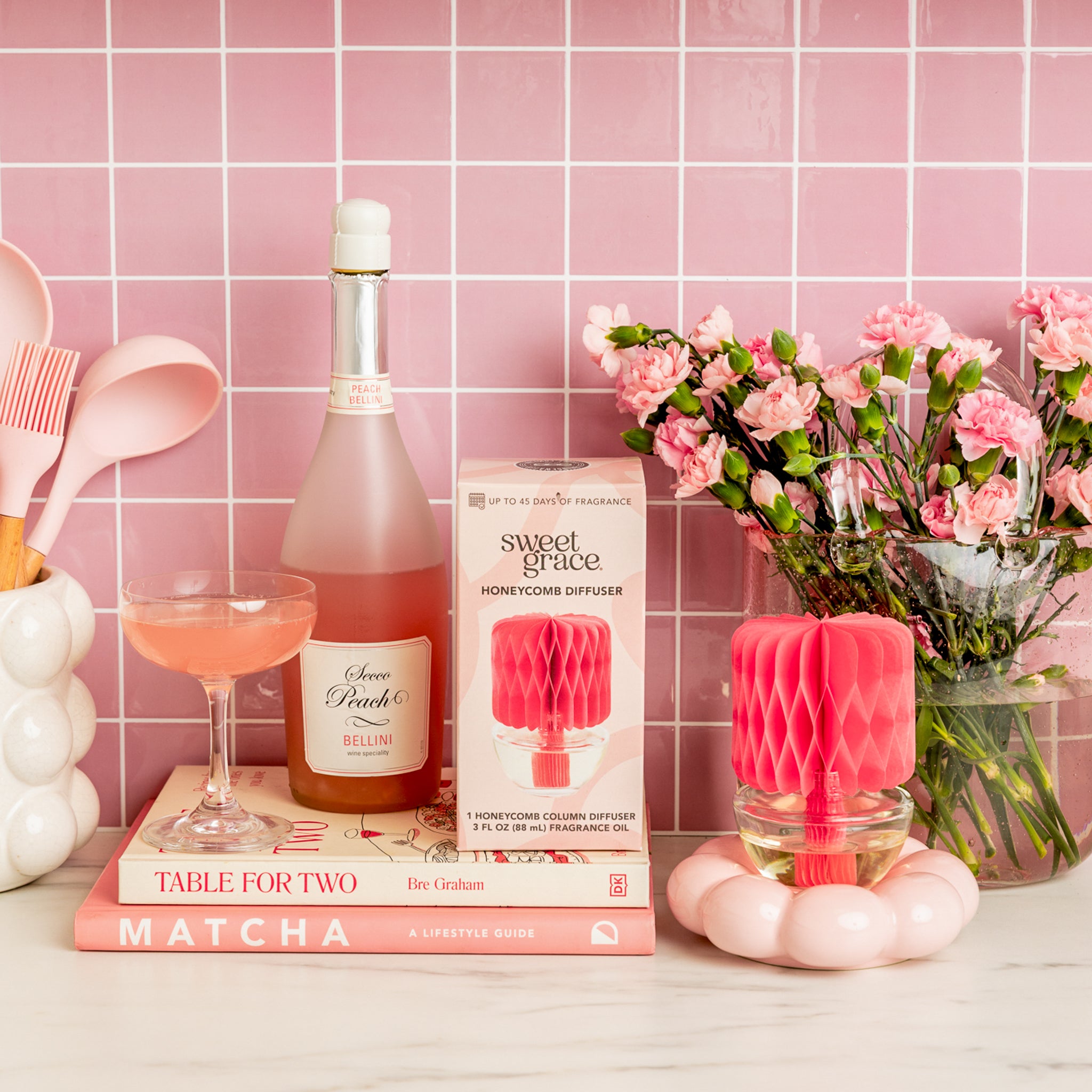 A pink-themed kitchen scene with pink utensils, a bottle and glass of pink Bellini, pink flowers, a Bridgewater Candle Sweet Grace Honeycomb Diffuser for lasting fragrance, and two pink books on a white counter against a pink tiled wall.