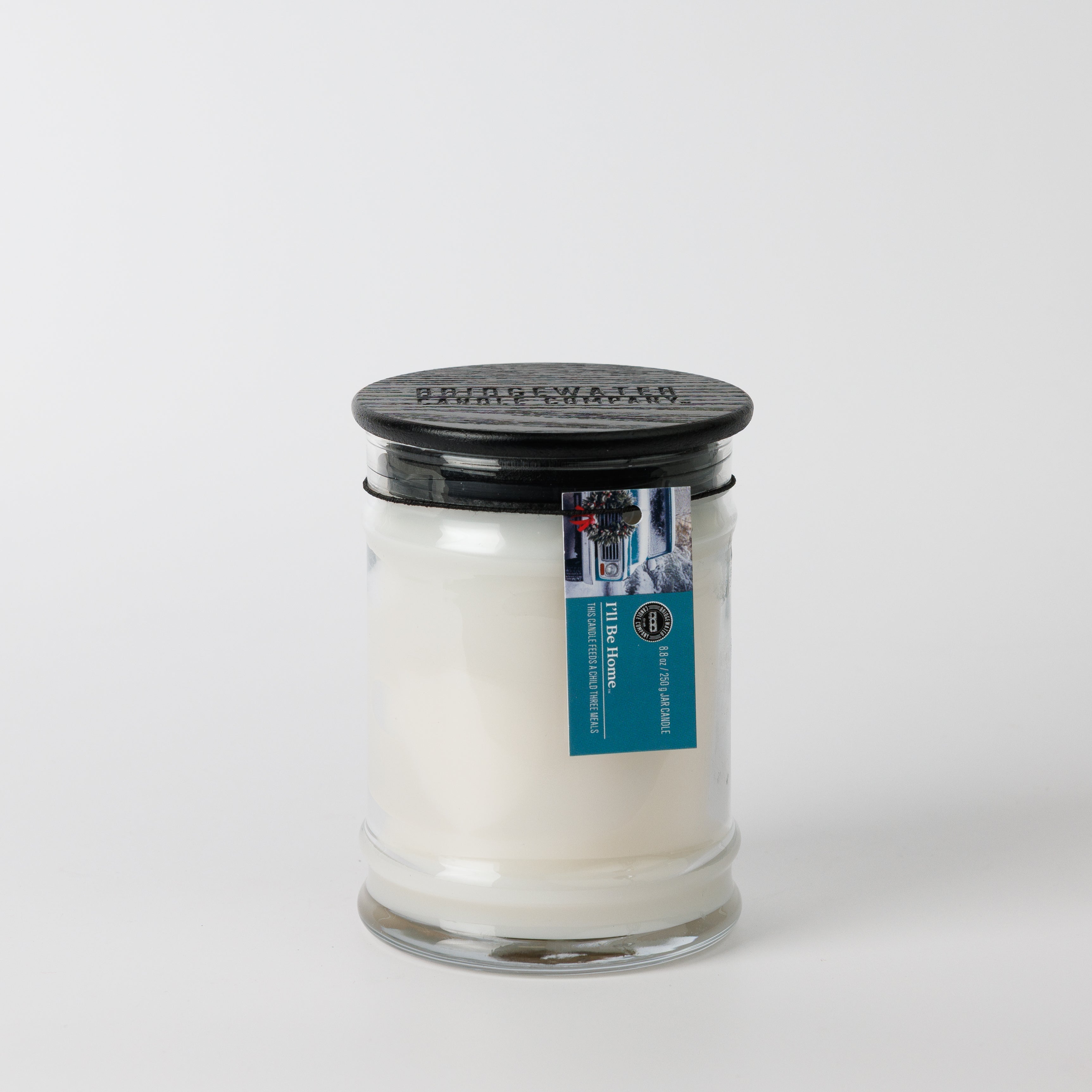 The Bridgewater Candle "I'll Be Home" 8oz jar candle features white soy-blend wax, a black lid, and a blue rectangular label with brand imagery on the neck. It stands against a white background.