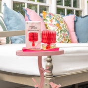A pink side table displays the Bridgewater Candle Sweet Grace Honeycomb Diffuser and box. A cushioned bench with colorful pillows sits nearby, filling the cozy space with continuous fragrance and charm near the window.