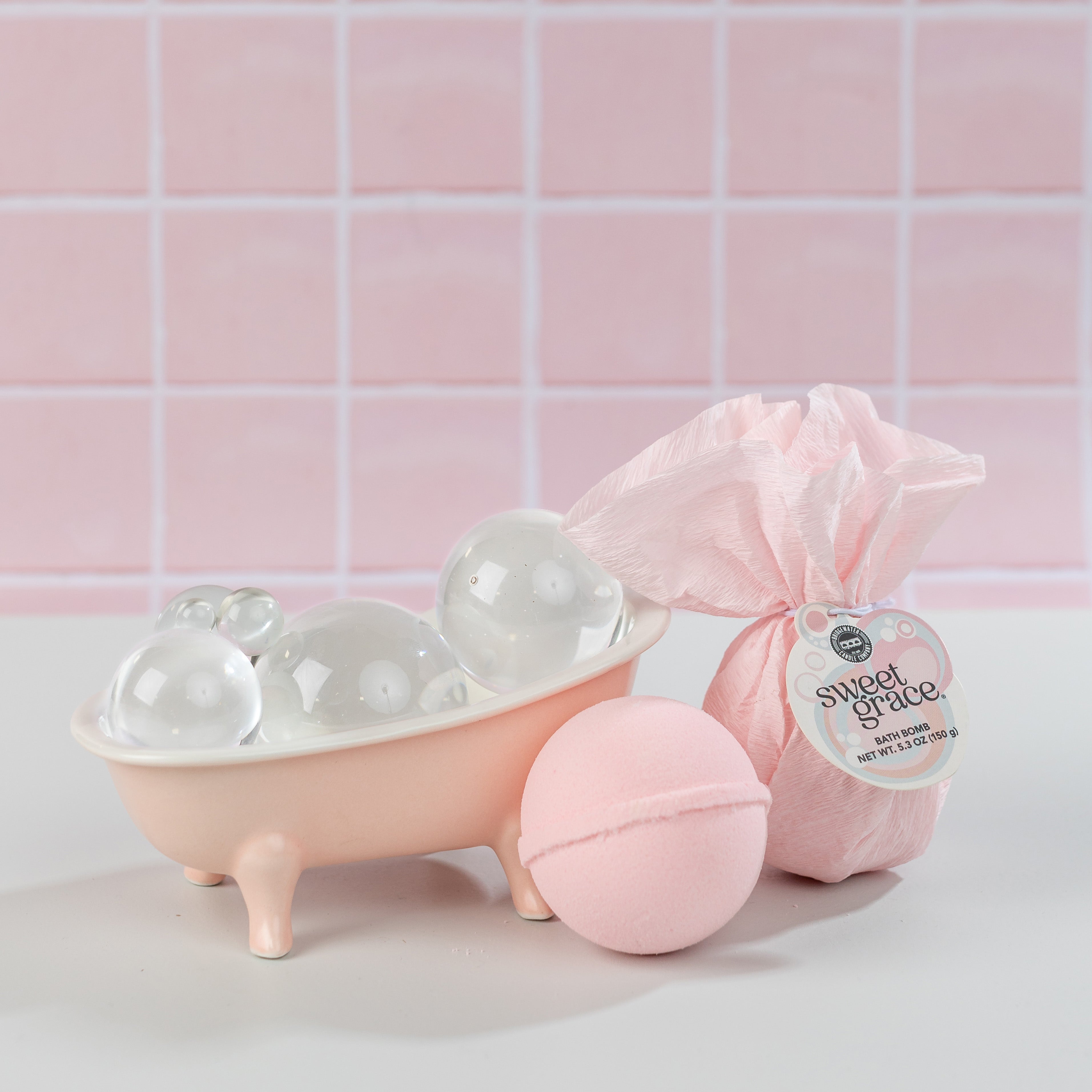 A small pink bathtub with bubbles sits beside Bridgewater Candles Bath Bomb 4 Pack–Sweet Grace and a pink-wrapped Sweet Grace product, all against a pink tiled background.