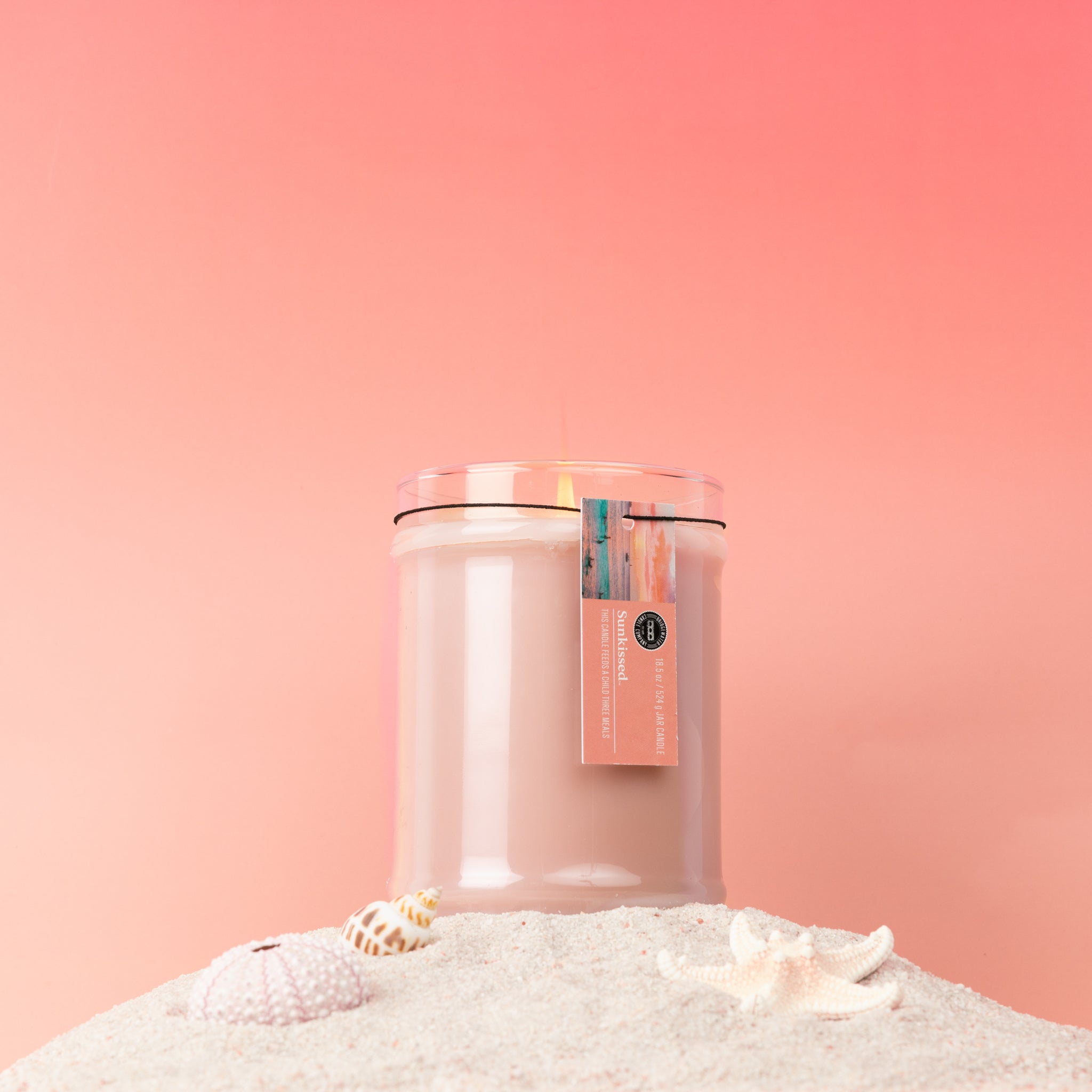 The Bridgewater Candle Sunkissed 18oz Large Jar Candle features soy-blend wax in a glass jar with a wooden lid and tag. Set on sand with seashells and a starfish, it creates a beachy vibe against a pink gradient background.