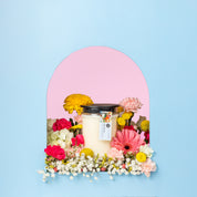 The Bridgewater Candle High Bloom 18oz Large Jar Candle, made with soy-blend wax and topped with a black lid, sits in a glass jar in an arched alcove surrounded by colorful flowers against a blue and pink background.