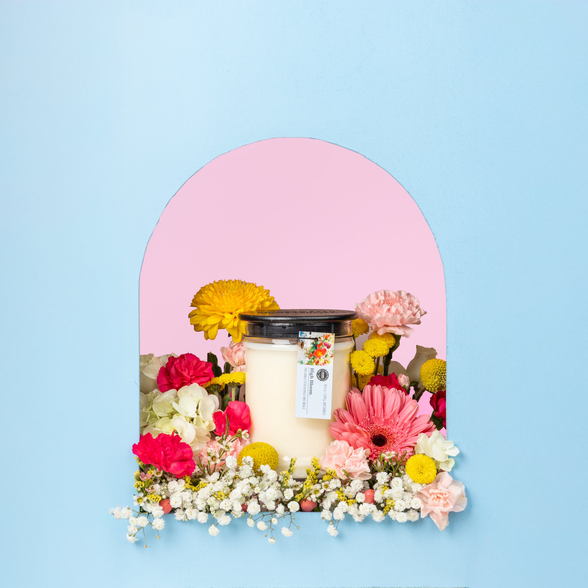 The Bridgewater Candle High Bloom 18oz Large Jar Candle, made with soy-blend wax and topped with a black lid, sits in a glass jar in an arched alcove surrounded by colorful flowers against a blue and pink background.
