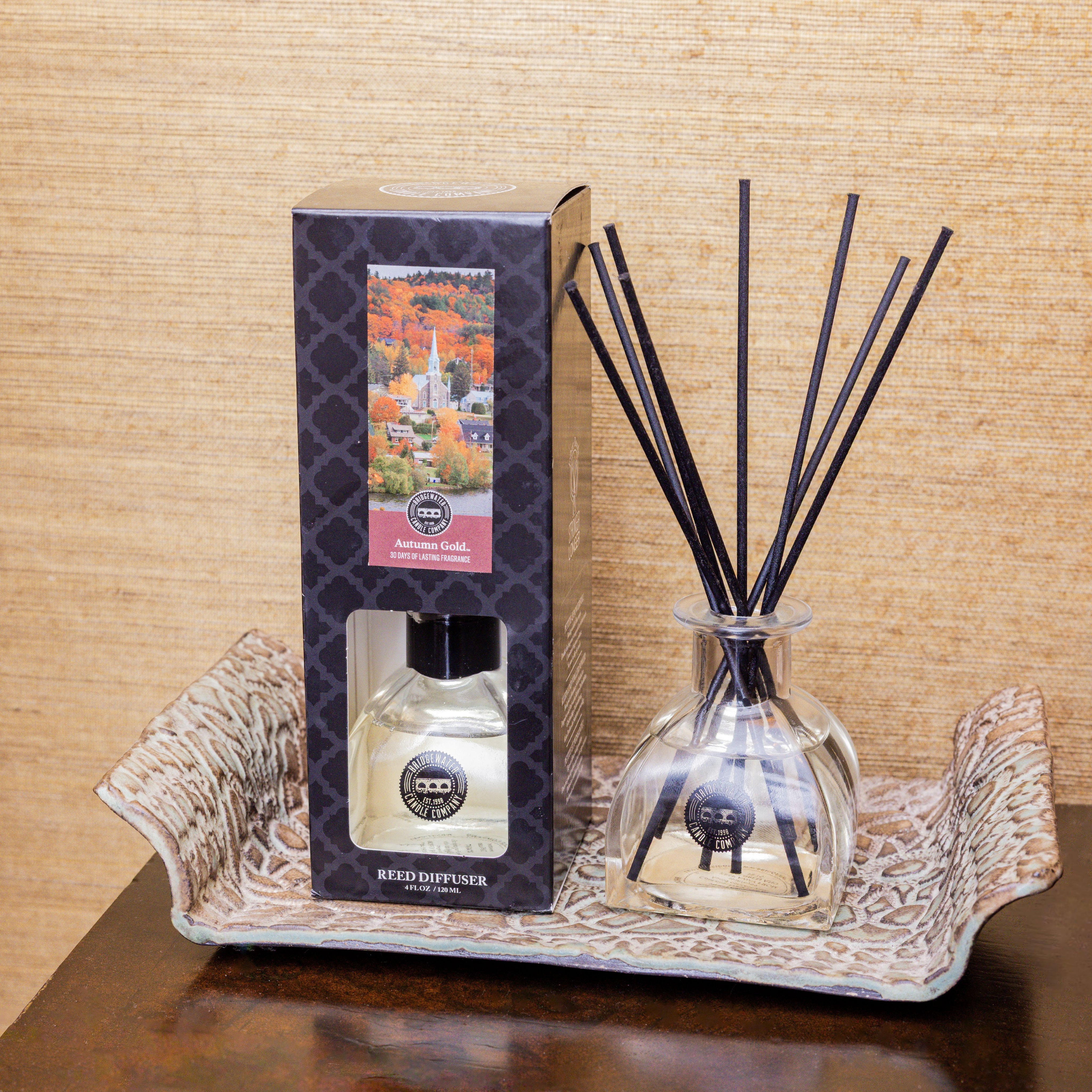 The Bridgewater Candle Autumn Gold Reed Diffuser with black sticks sits on a patterned tray beside its decorative box, showcasing a scenic autumn landscape and filling your space with an inviting, cozy fall fragrance.