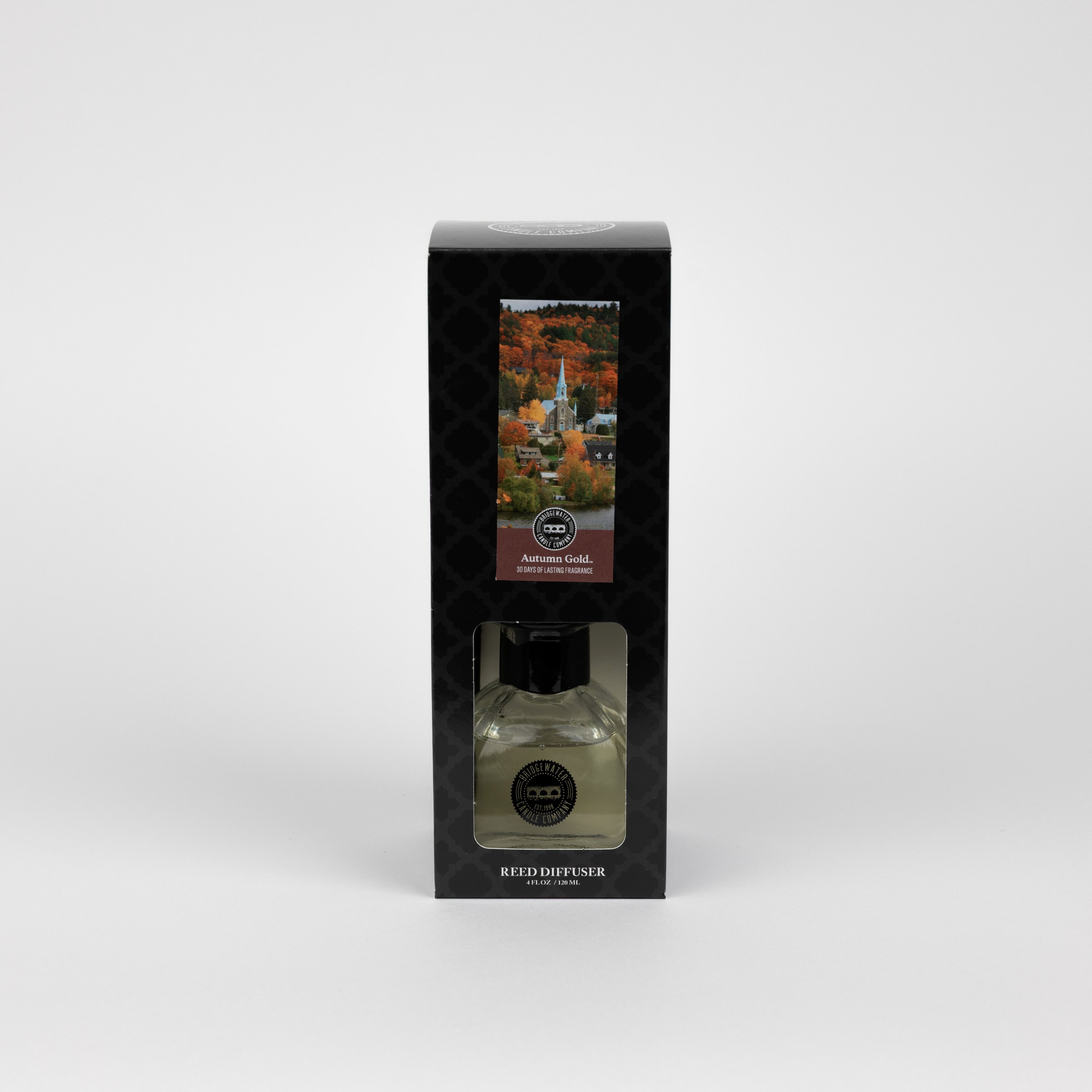 The Bridgewater Candle Autumn Gold Reed Diffuser comes in a black box with a clear window, showing the bottle inside. The box features autumn trees and a white building, making it the perfect seasonal décor for your home.