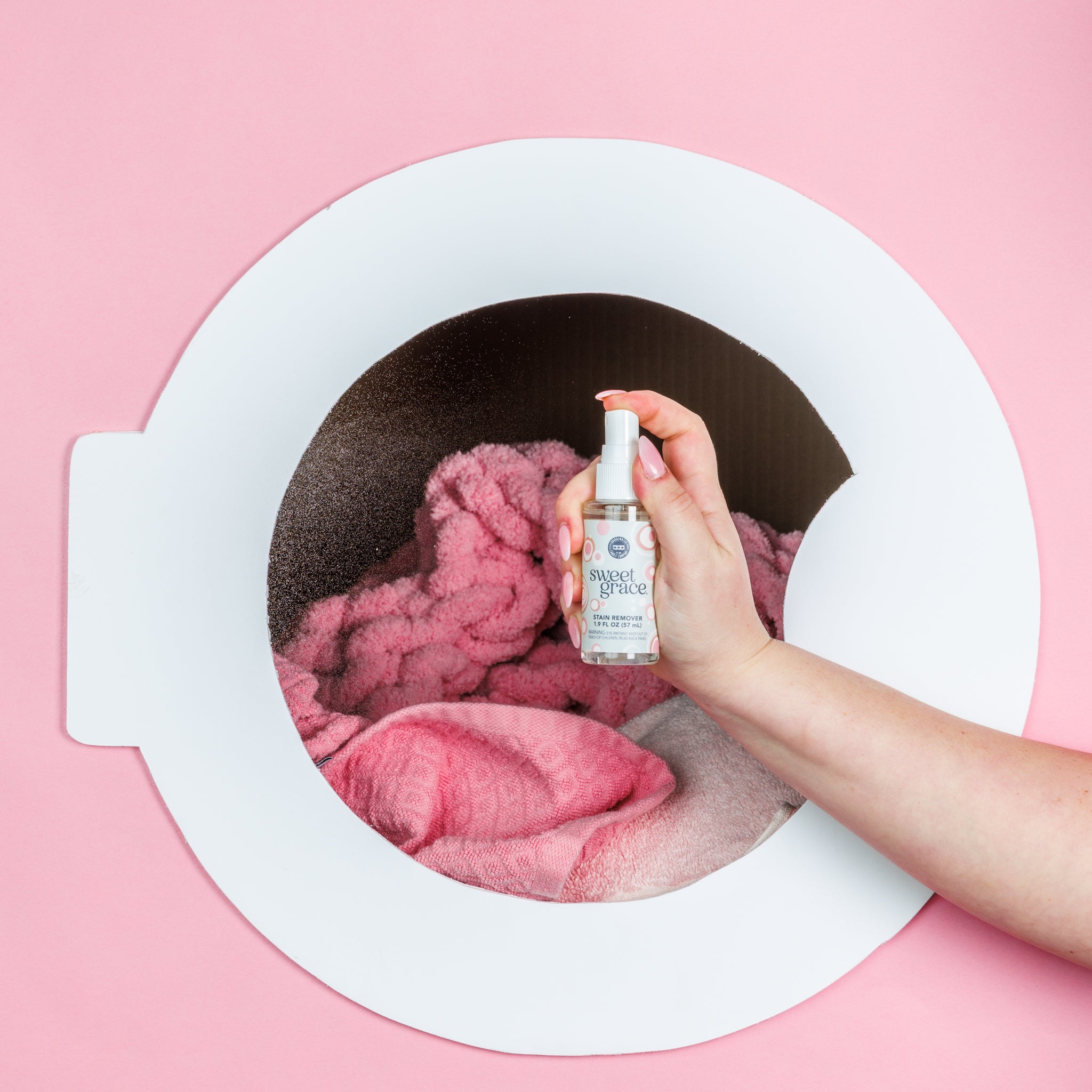 A hand holds a Bridgewater Candle Sweet Grace Stain Remover - Travel Size spray bottle over pink towels inside a white front-loading washing machine, set against a pink background.