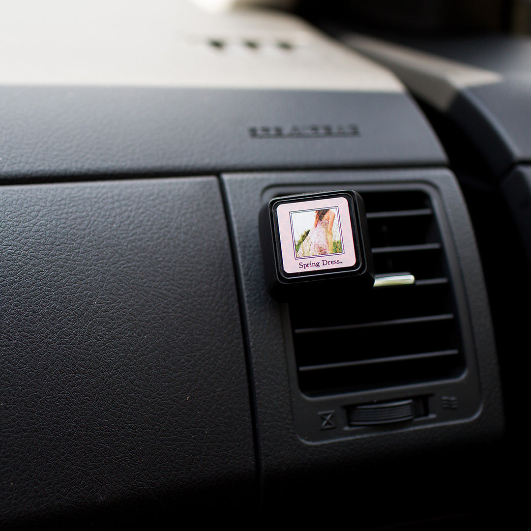 A square Bridgewater Candle Auto Vent Clip-Spring Dress attached to a vehicle dashboard, displaying an image of a person in a floral dress with the text Spring Dress below it.
