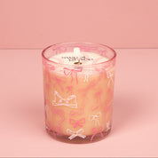 The Bridgewater Candle Sweet Grace Pink Bow Candle, made with soy-blend wax, sits in a glass holder decorated with pink and white bows on a pink surface, matching the background. The candle label displays "Sweet Grace.