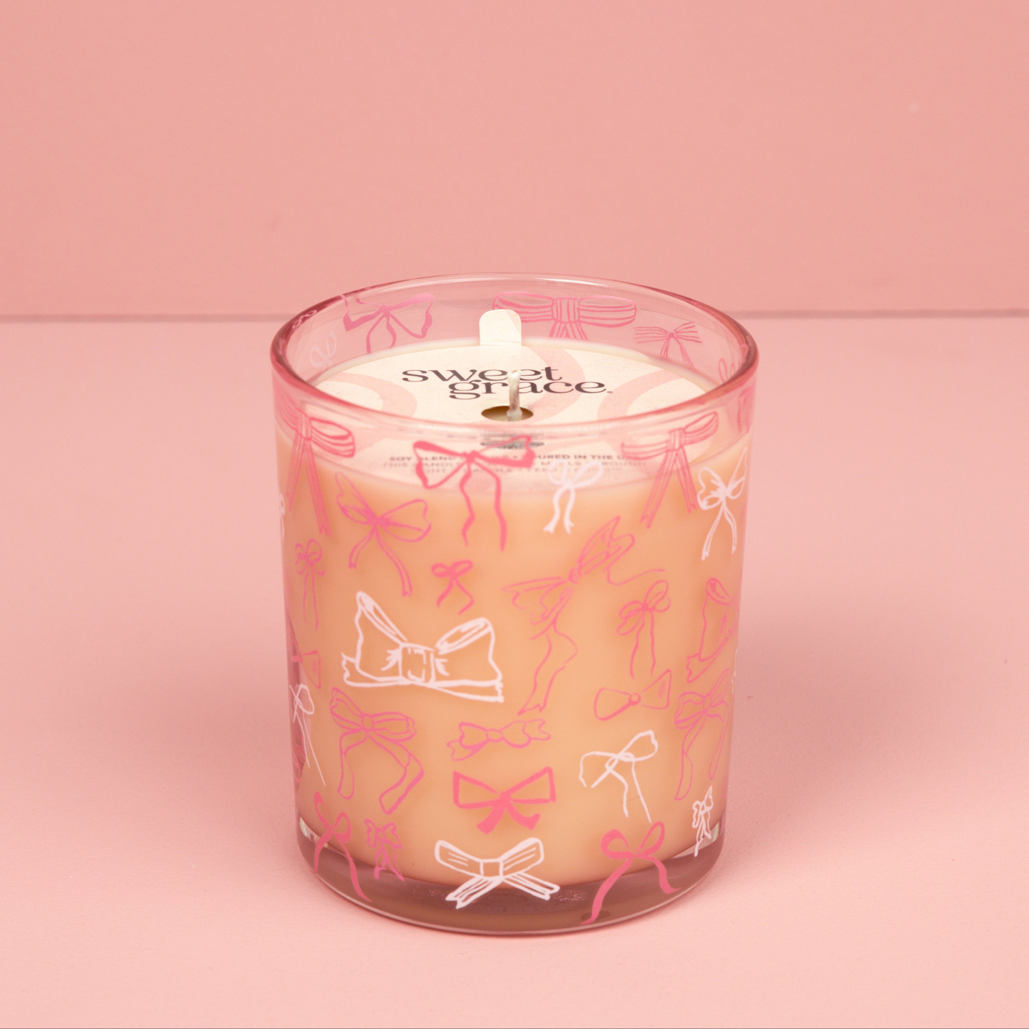 The Bridgewater Candle Sweet Grace Pink Bow Candle, made with soy-blend wax, sits in a glass holder decorated with pink and white bows on a pink surface, matching the background. The candle label displays "Sweet Grace.