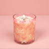 The Bridgewater Candle Sweet Grace Pink Bow Candle, made with soy-blend wax, sits in a glass holder decorated with pink and white bows on a pink surface, matching the background. The candle label displays "Sweet Grace.