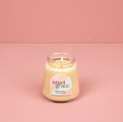 A lit Bridgewater Candle Sweet Grace Collection Candle #078, made with soy-blend wax, glows in a petite glass jar on a pink surface with a matching pink background.