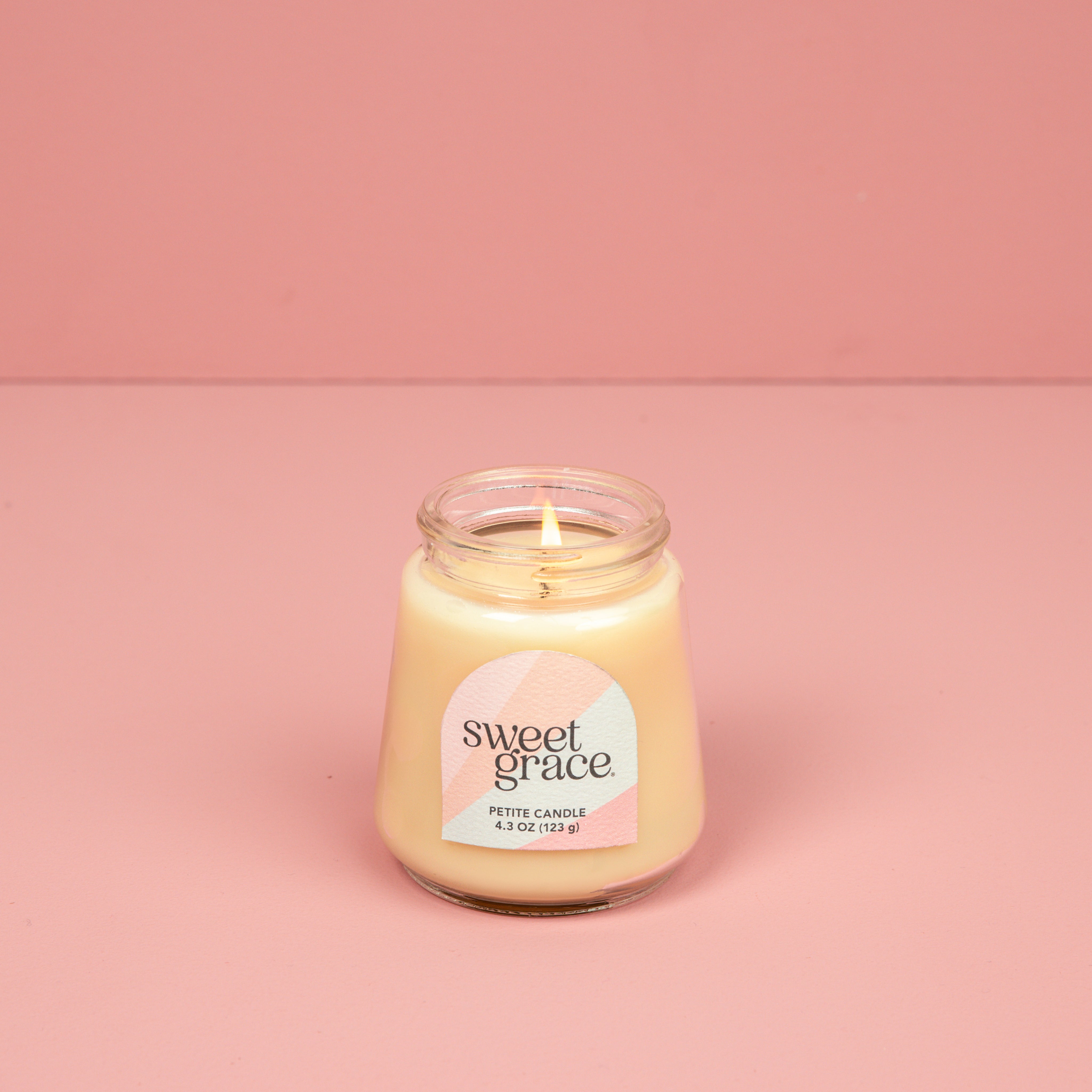 A lit Bridgewater Candle Sweet Grace Collection Candle #078, made with soy-blend wax, glows in a petite glass jar on a pink surface with a matching pink background.