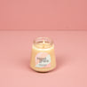 A lit Bridgewater Candle Sweet Grace Collection Candle #078, made with soy-blend wax, glows in a petite glass jar on a pink surface with a matching pink background.