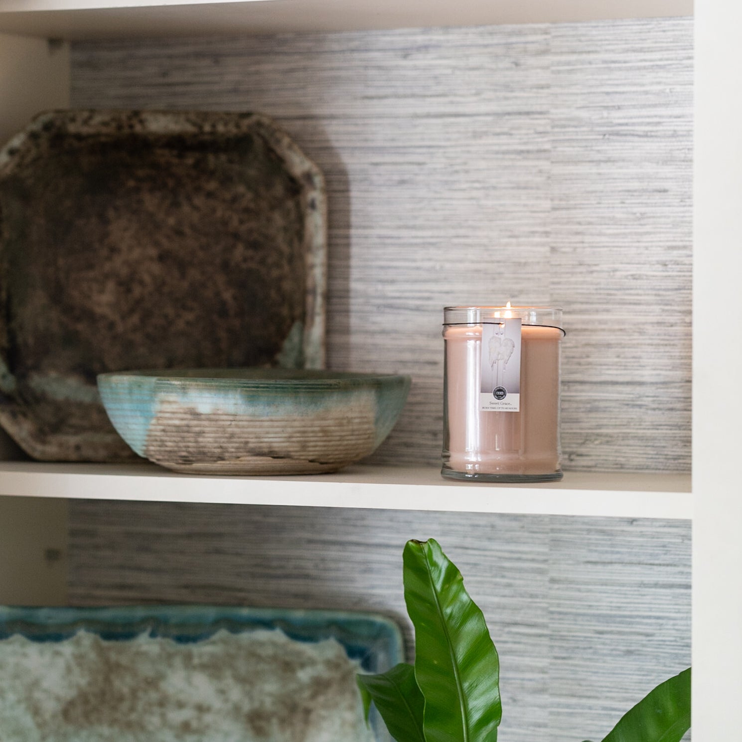 A Bridgewater Candle Sweet Grace 18oz Large Jar Candle glows on a shelf next to two ceramic bowls, with a green plant leaf visible at the bottom right against a textured light gray background.