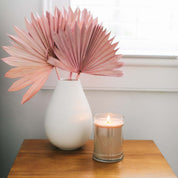 A white vase with pink dried palm leaves and a lit Bridgewater Candle Sweet Grace 18oz Large Jar Candle sit on a wooden table in front of a window with white blinds.