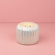 A white, iridescent ceramic holder with vertical grooves cradles a lit Bridgewater Candle Sweet Grace Collection Candle #071, a double-wick soy-blend promising long-lasting fragrance. Displayed on a pink surface against a pink background.