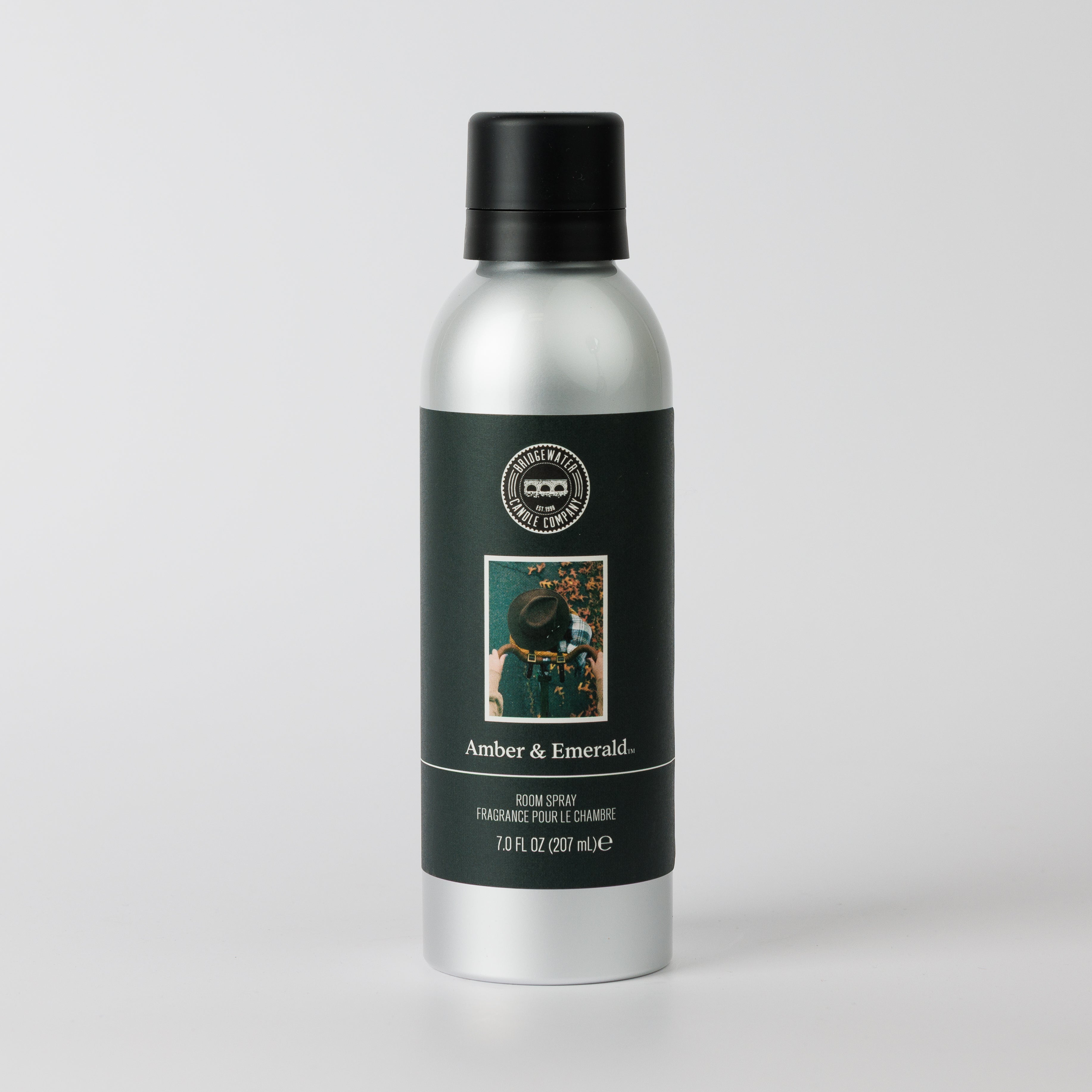 A silver spray bottle with a black cap and dark green label reading "Amber & Emerald Non-Aerosol Room Spray" by Bridgewater Candle features a small image of a person holding flowers, shown against a plain white background.