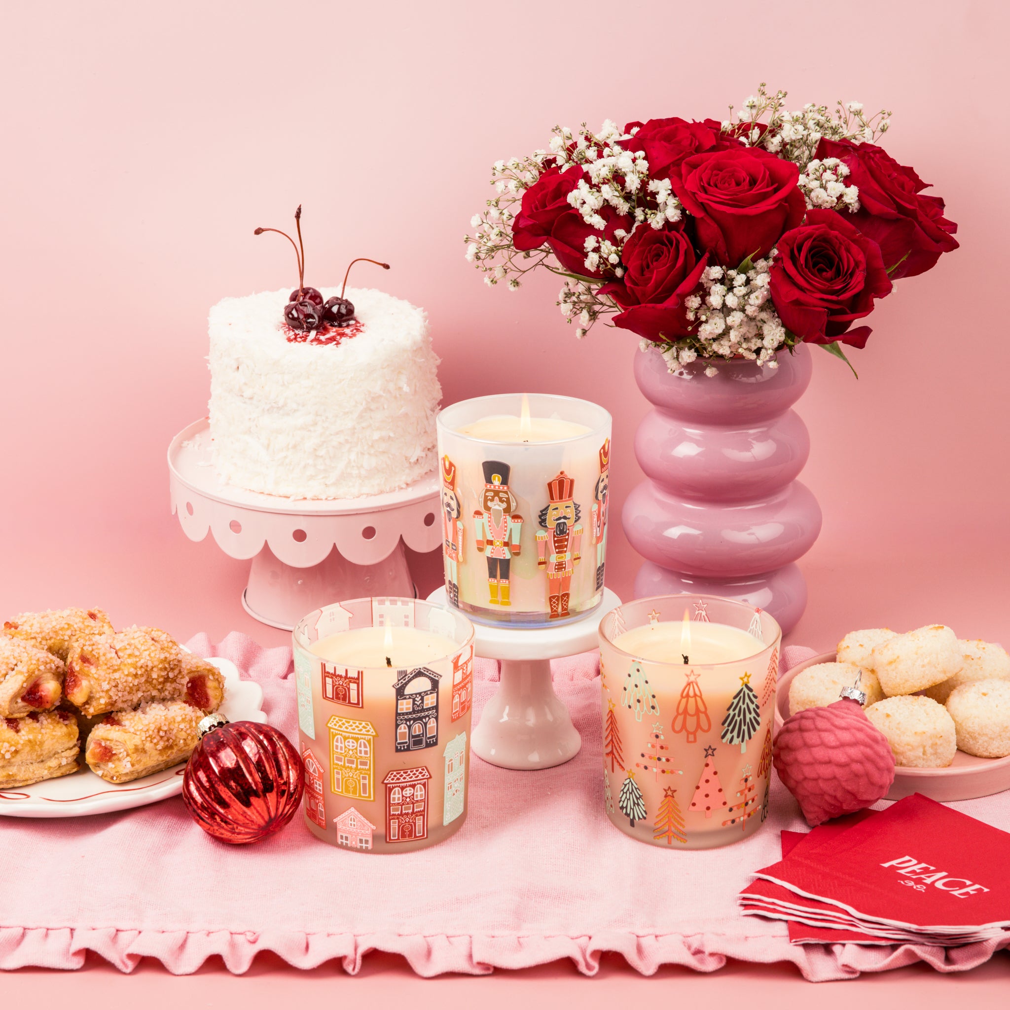 A festive table with a coconut cake topped with cherries, a bouquet of red roses, three Bridgewater Candle Sweet Grace Holiday Nutcracker Candles, Christmas ornaments, pastries, donuts, and red Peace napkins on a pink scalloped tablecloth.
