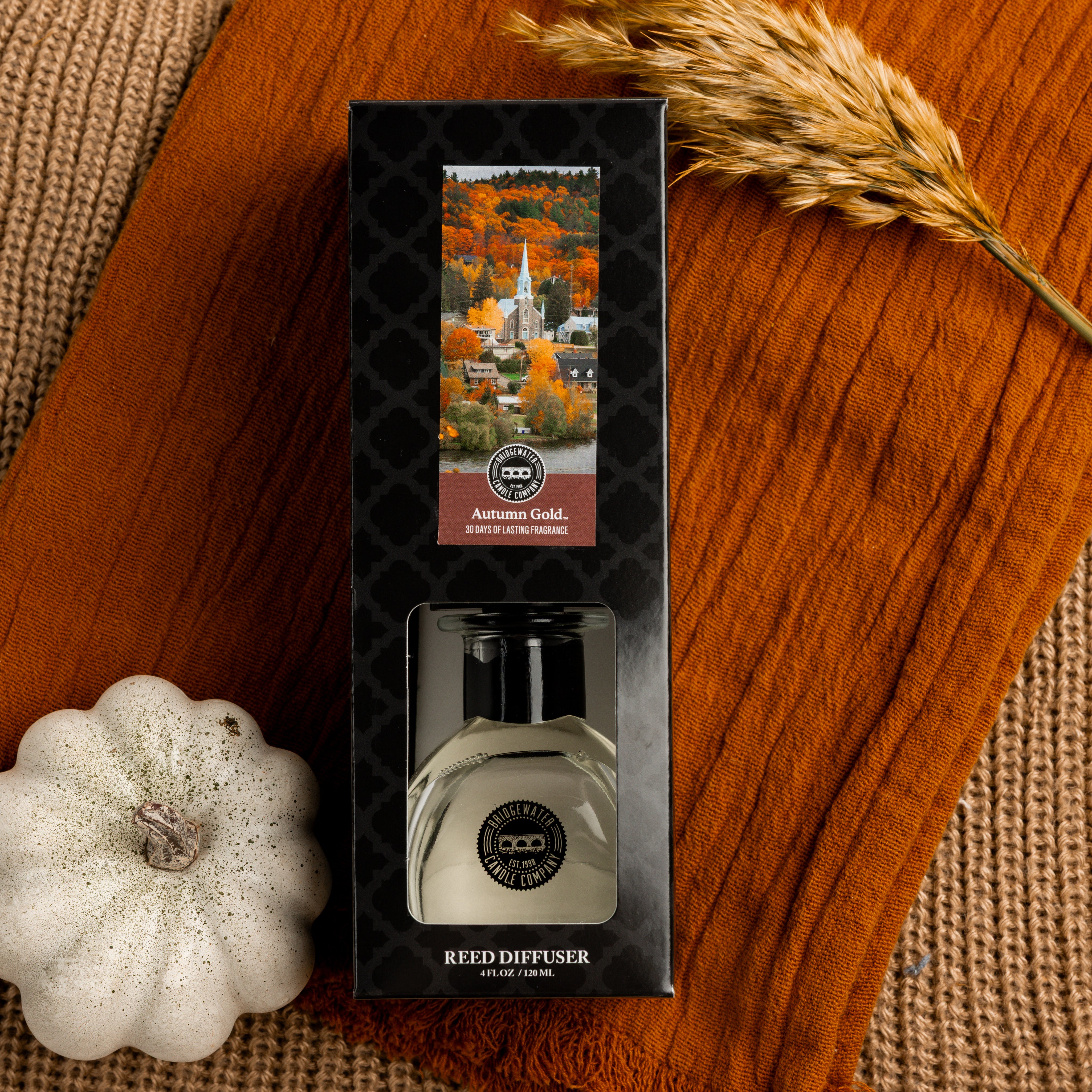 The Bridgewater Candle Autumn Gold Reed Diffuser comes in fall foliage packaging and sits on orange knit fabric beside a white pumpkin and dried wheat, bringing autumn fragrance and charm to your home décor.
