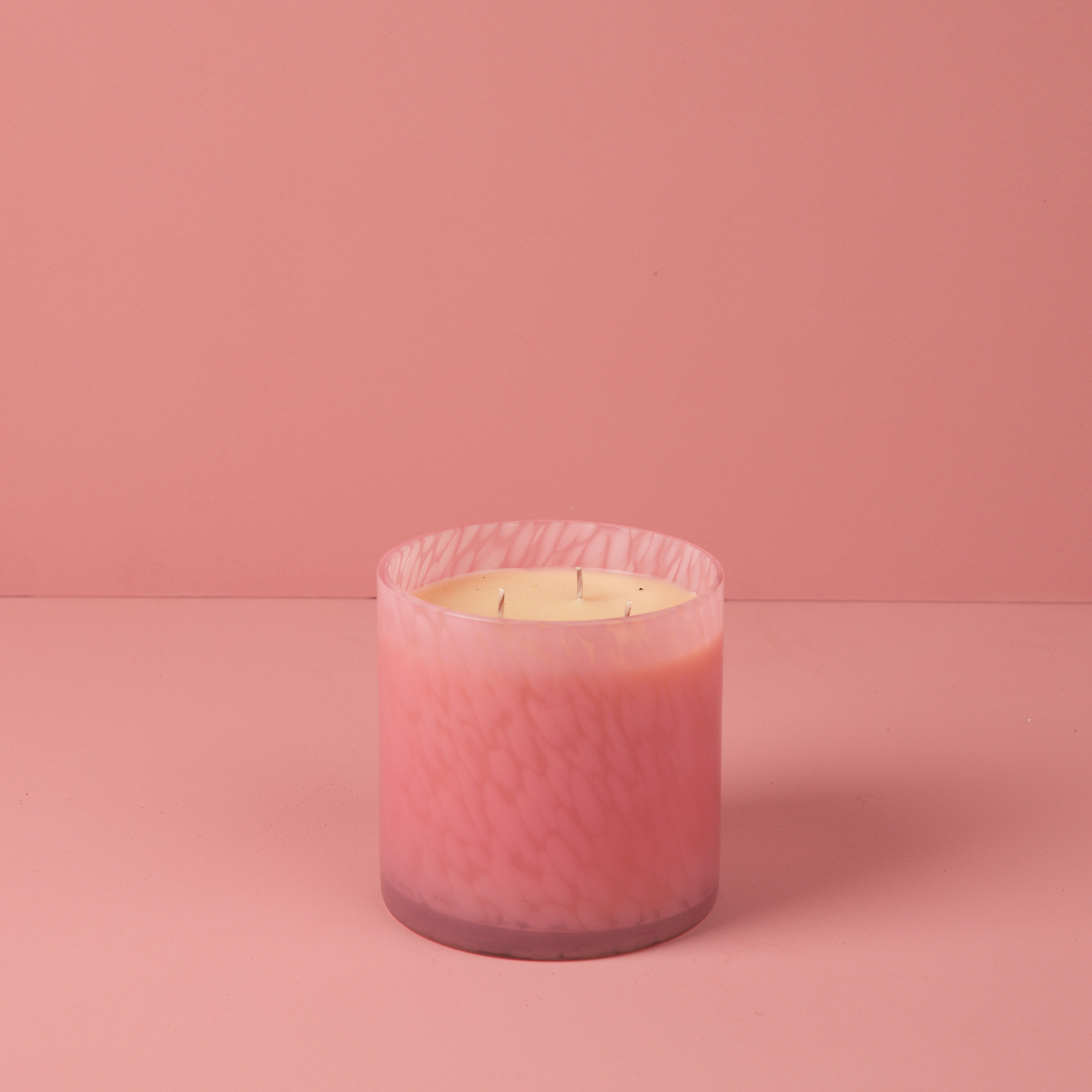 The Bridgewater Candle Sweet Grace Collection Candle #073, a pink marbled glass three-wick candle made from soy-blend wax, rests on a smooth pink surface with a matching pink background.